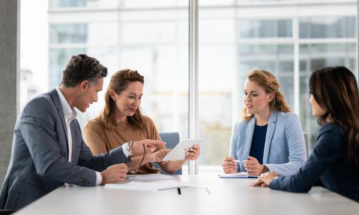 A man and three women in business outfits sit in a meeting room and talk to each other