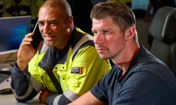 Two men in a control room, one wearing a high-visibility jacket and talking on the phone, while the other looks intently at a monitor.