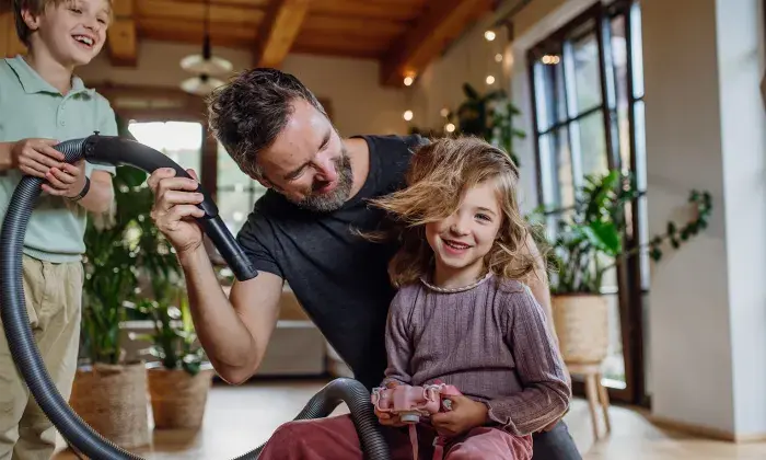 A father playfully using a vacuum hose to blow air into his daughter's hair while his son laughs nearby, creating a fun family moment indoors.