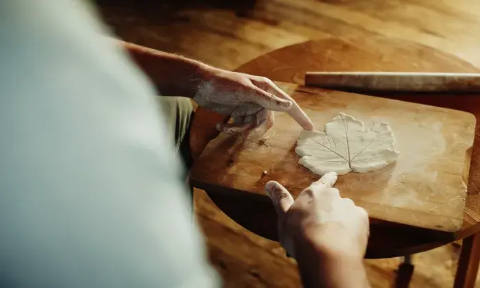 A person with clay-covered hands shapes a leaf pattern out of clay on a wooden board. The scene captures a moment of careful craftsmanship and creativity in a warm, natural setting.