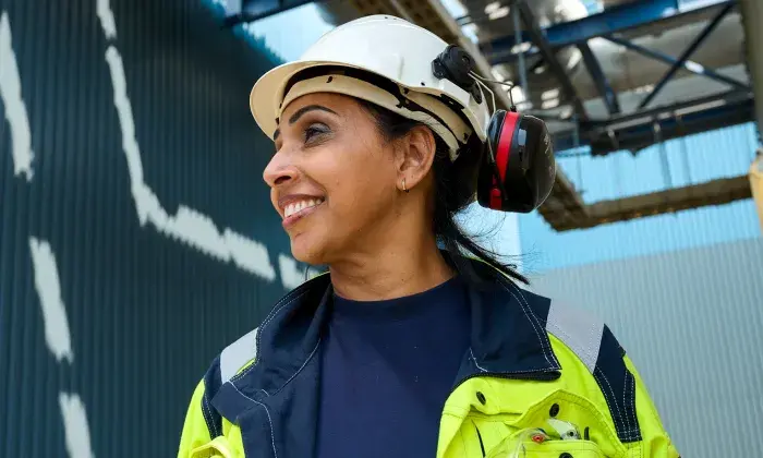 A worker wearing a hard hat and ear protection, smiling while standing outdoors at an industrial site.
