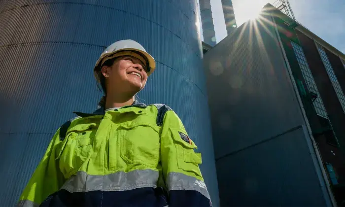 A worker in a high-visibility jacket and hard hat smiling while standing outside near industrial silos, with sunlight shining behind them.