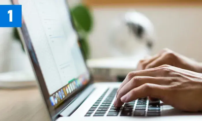 Close-up of hands typing on a laptop keyboard, with a document open on the screen and a blue box with the number '1' in the top left corner.