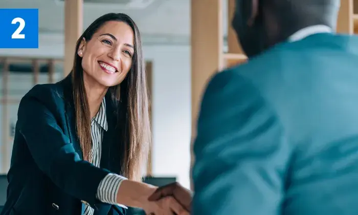 A smiling woman in a professional setting shaking hands with another person, with a blue box containing the number '2' in the top left corner.