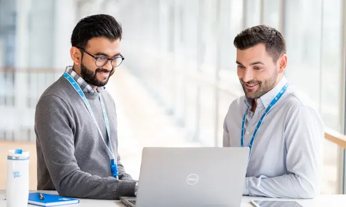 Two male colleagues sitting at a table, smiling while working together on a laptop in a bright office space, with a notebook and a Uniper-branded water bottle on the table