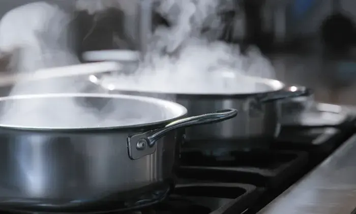 Two stainless steel pots on a stovetop, with steam rising from them, indicating that the contents are being heated or boiled.