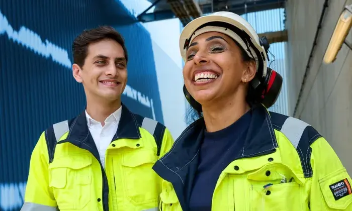 Two workers in safety gear, one wearing a helmet and ear protection, smiling while standing in an industrial setting.