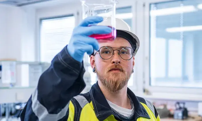 A technician in safety gear and a hard hat holds up a beaker with a pink liquid, carefully inspecting it in a laboratory setting.
