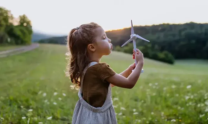 A young girl standing in a field at sunset, holding a toy wind turbine and blowing on it, with trees and hills in the background.