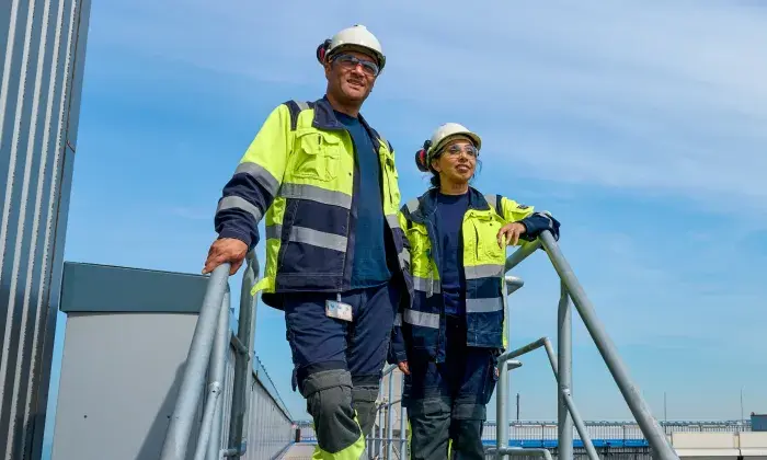 Two workers in high-visibility safety jackets, hard hats, and protective glasses standing on an outdoor industrial platform under a clear sky.