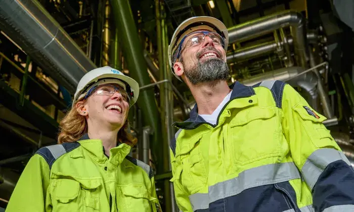 Two workers in high-visibility safety jackets, hard hats, and protective glasses smiling while standing in an industrial setting, with metal pipes and machinery in the background.