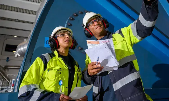 Two industrial workers in high-visibility jackets, hard hats, and ear protection reviewing documents and taking notes while inspecting machinery inside a facility.