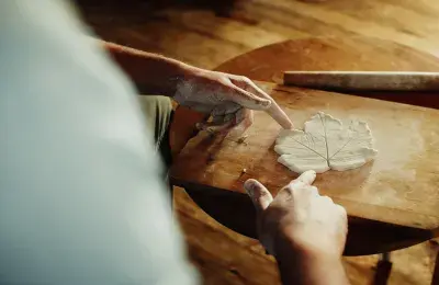 A person with clay-covered hands shapes a leaf pattern out of clay on a wooden board. The scene captures a moment of careful craftsmanship and creativity in a warm, natural setting.
