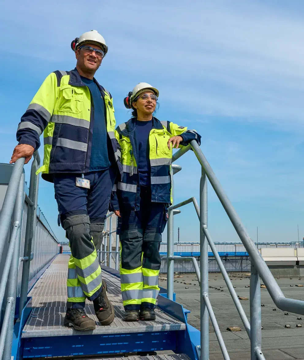 Two workers in safety gear stand on a metal staircase overlooking an industrial rooftop with a clear sky in the background.
