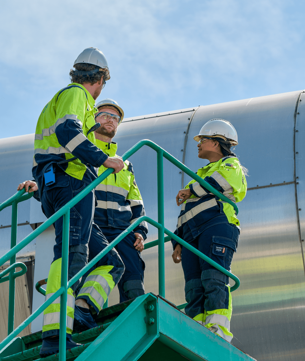 Three industrial workers in protective gear standing on an outdoor staircase, discussing work at an energy facility