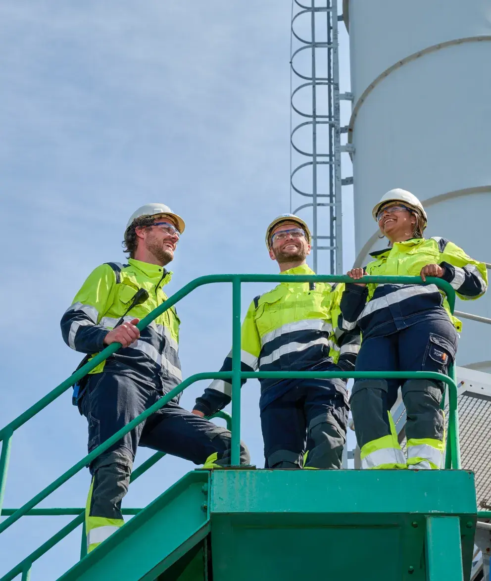 Three workers in safety helmets and high-visibility clothing standing on a green metal staircase, smiling and looking out at an industrial site.