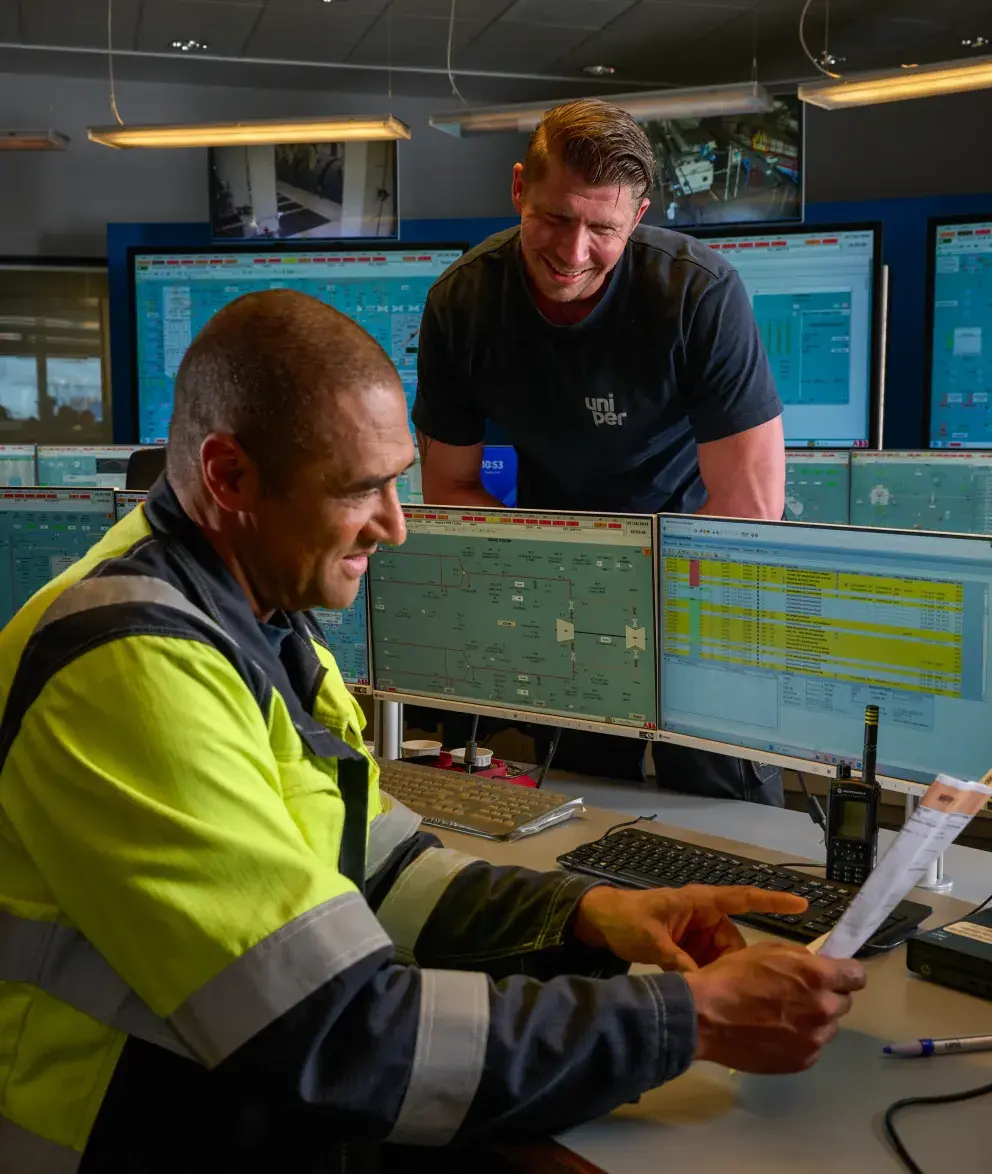 Two workers in a control room, one seated and reviewing a document, while the other stands next to him, smiling, with multiple monitors displaying technical data in the background.