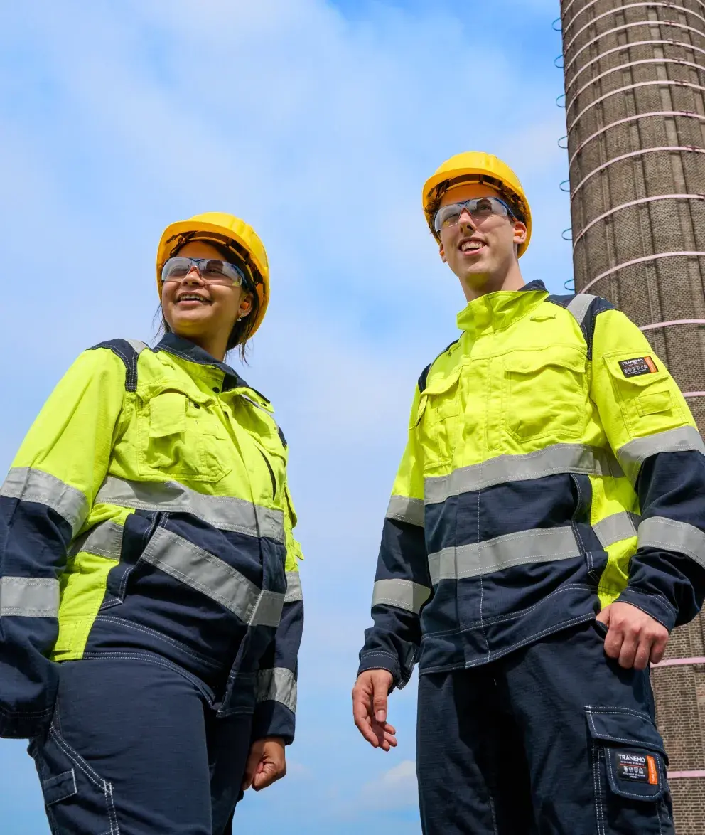 Two industrial workers in high-visibility jackets, hard hats, and safety glasses standing outdoors under a blue sky near a tall industrial structure.
