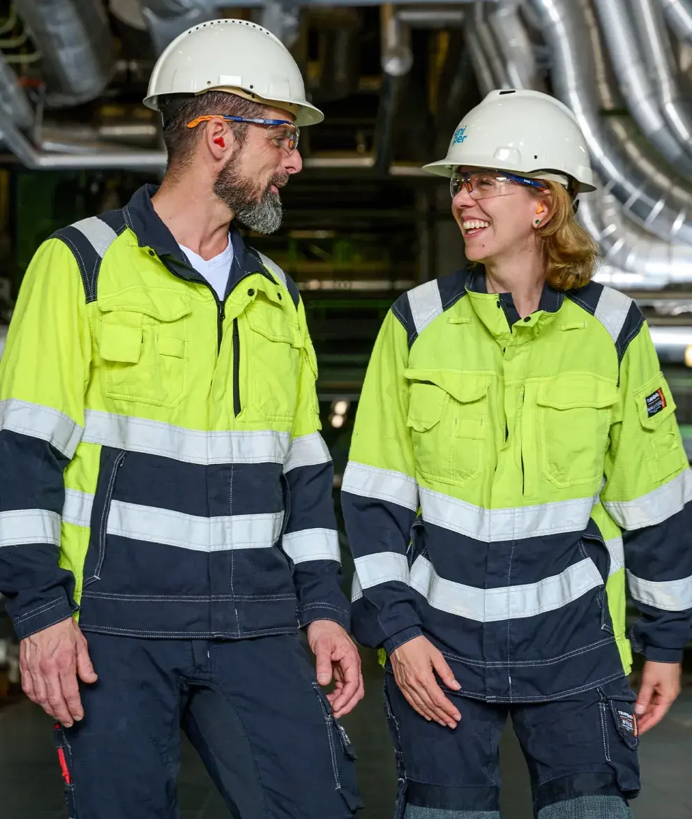 Two Uniper workers in high-visibility clothing and safety helmets smiling and walking together inside an industrial facility with metal piping in the background.