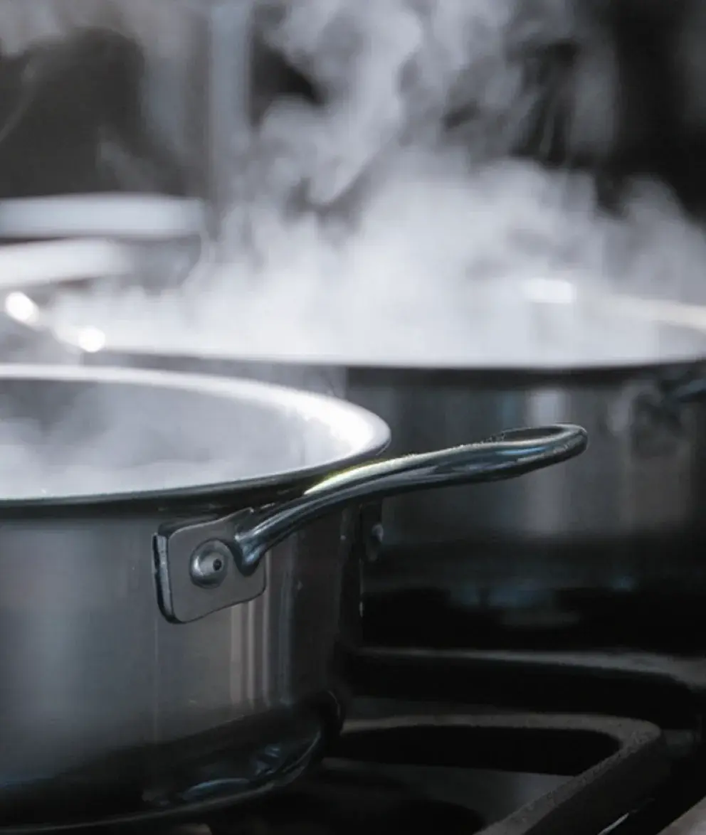 Two stainless steel pots on a stovetop, with steam rising from them, indicating that the contents are being heated or boiled.