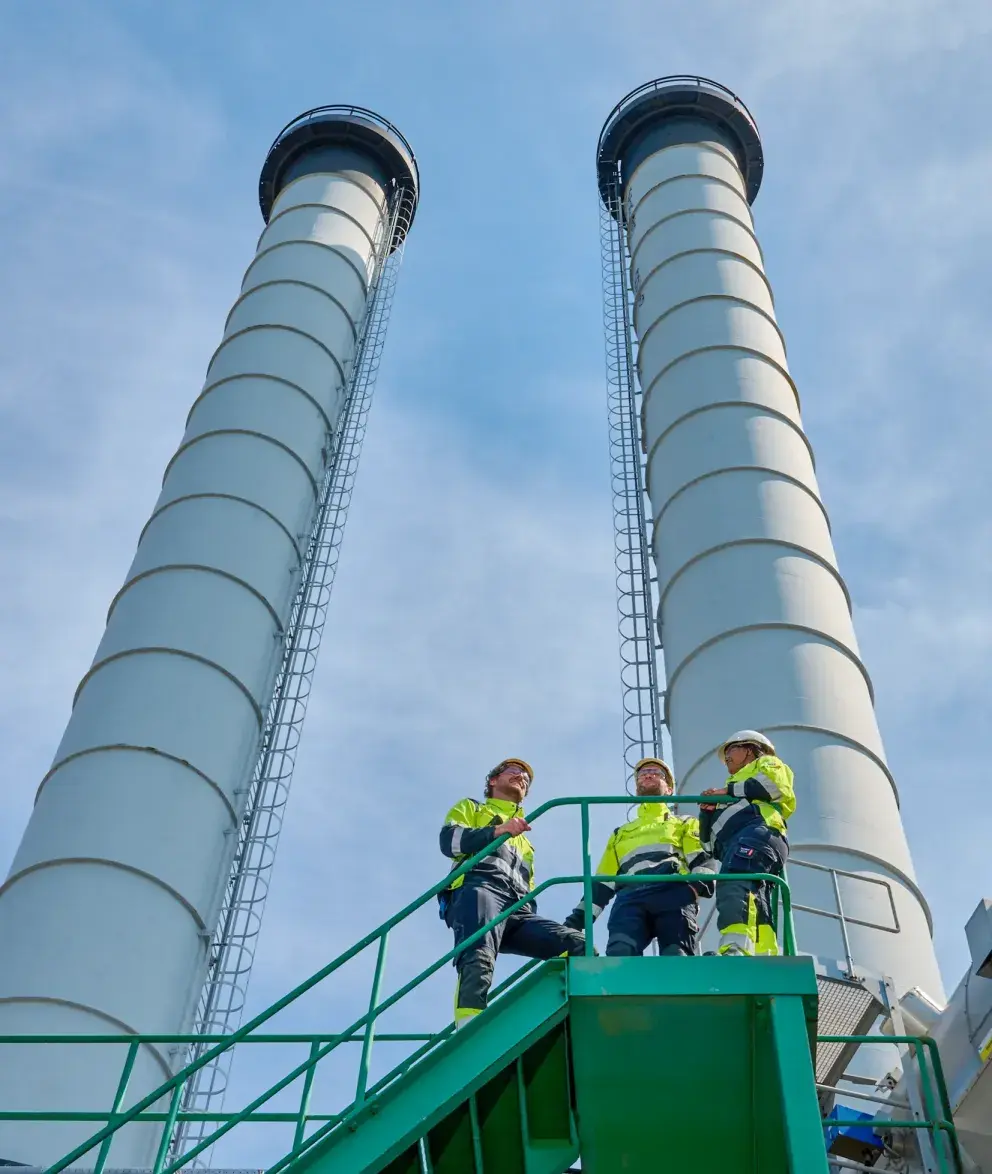 Three workers in high-visibility gear stand on a green staircase in front of two large industrial chimneys under a clear blue sky.