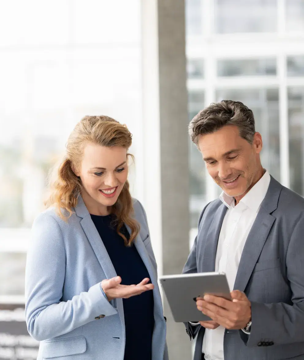 Two professionals in business attire discussing something on a tablet, smiling in a bright office environment.