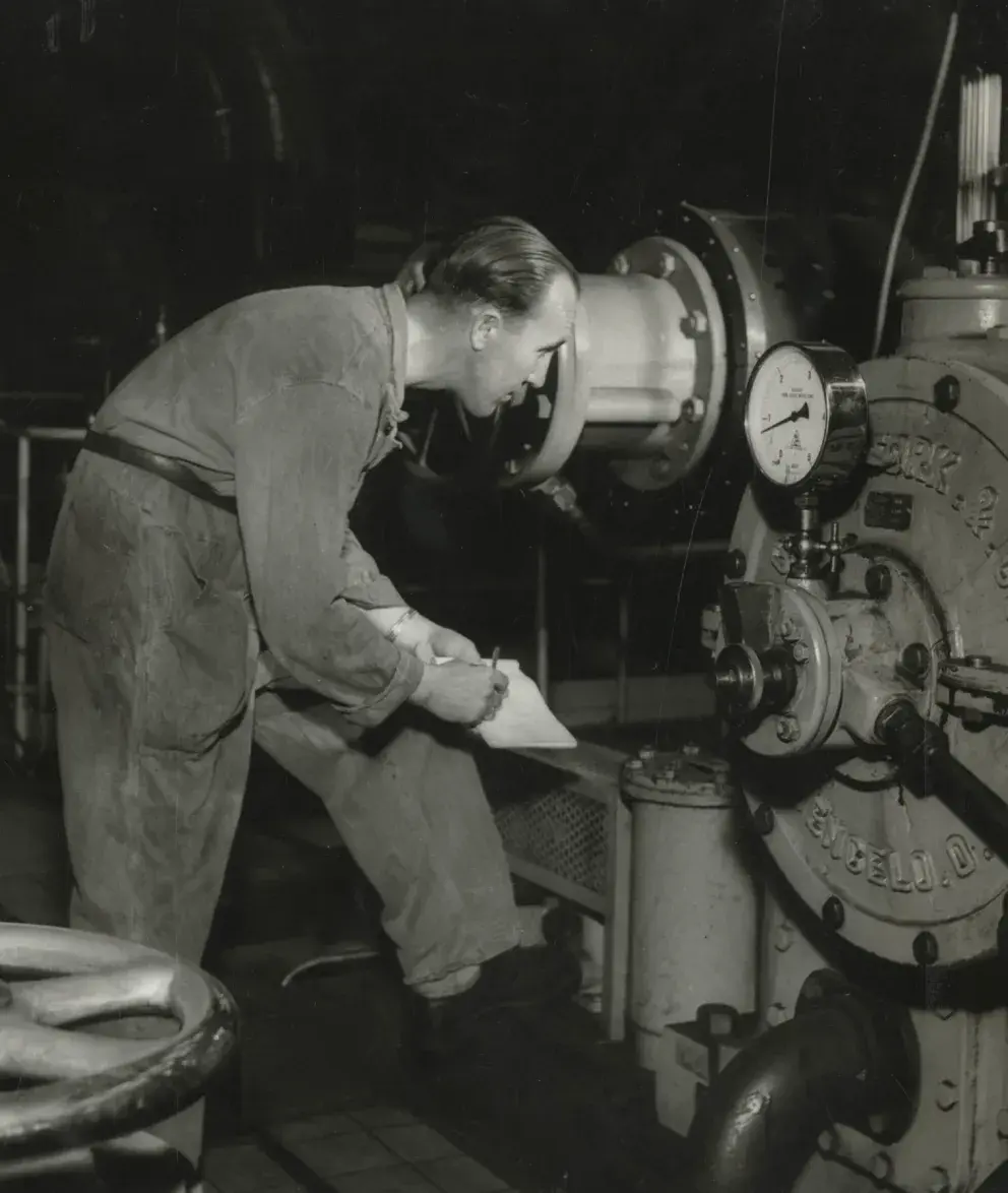 A historical black-and-white photo of a worker in a factory, checking a machine and taking notes.