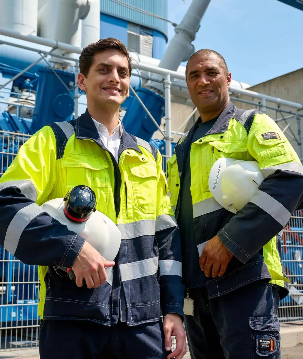 Two Uniper employees wearing high-visibility jackets and holding safety helmets, standing in front of industrial equipment at a power plant.