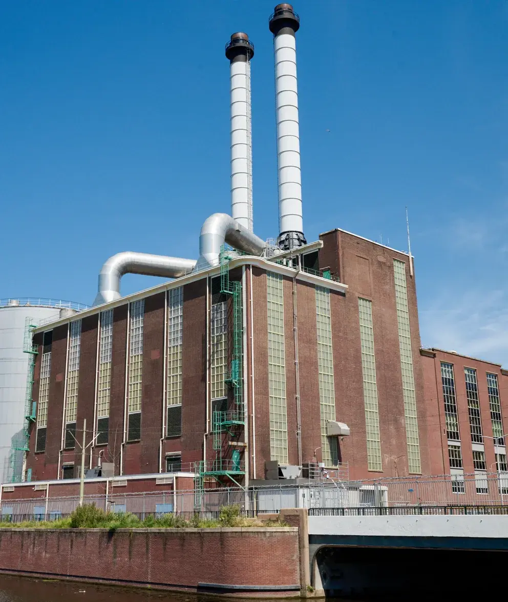 Exterior view of a brick-built city power plant in The Hague, featuring tall chimneys and industrial pipes under a clear blue sky.