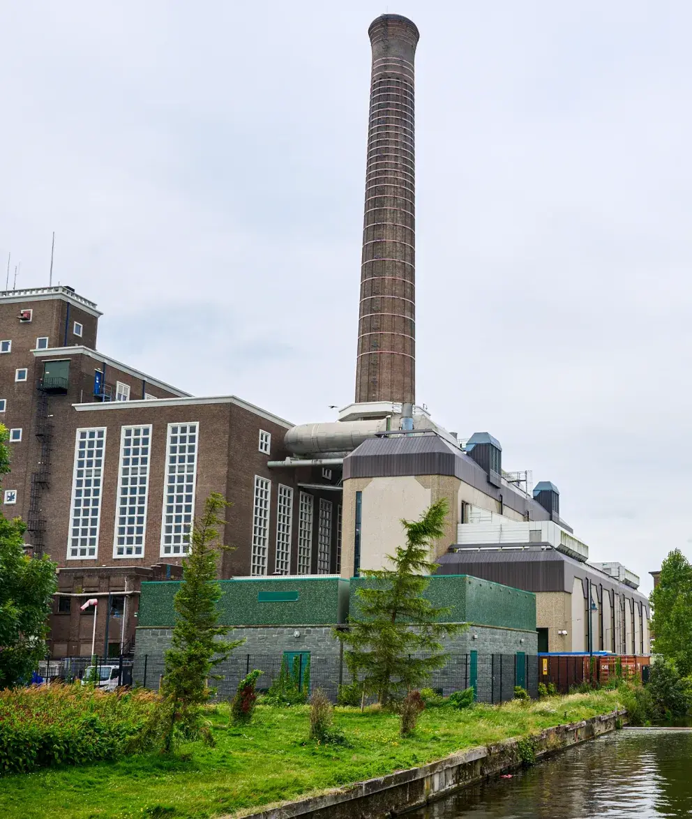 Exterior view of the Uniper city power plant in Leiden, featuring a tall brick chimney and industrial buildings near a canal with green surroundings