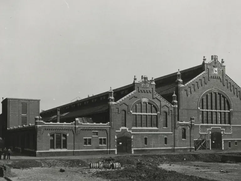 A historical black-and-white photo of a large brick factory building with a distinctive tall, patterned chimney and several workers standing in front of it.