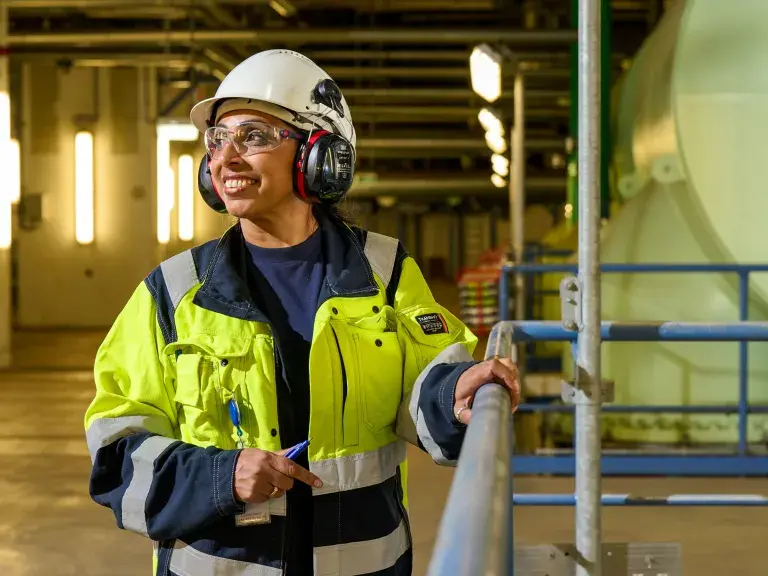 Worker wearing a yellow safety jacket, helmet, ear protection, and safety glasses, standing inside an industrial facility and looking to the side with a smile