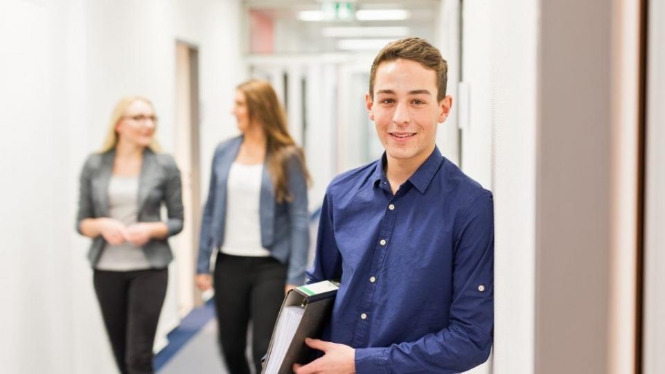 Männlicher Uniper-Auszubildender mit einem Aktenordner und zwei weiblichen Mitarbeitern im Hintergrund