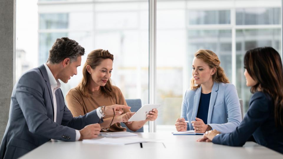 Ein Mann und drei Frauen in Business Kleidung sitzen in einem Besprechungsraum und reden miteinander