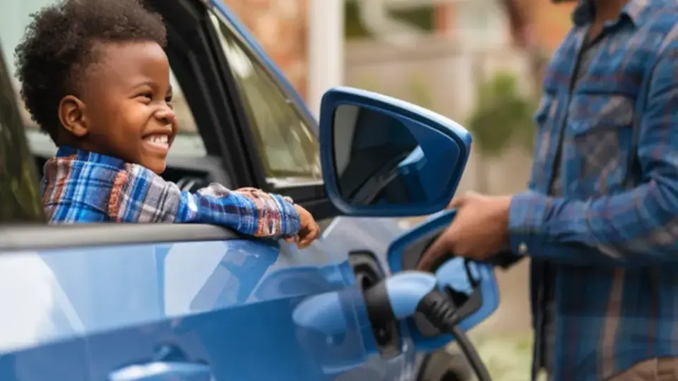The image shows a heartwarming scene of a man and a young child near a blue electric car. The man, wearing a plaid shirt, is smiling and engaged in the activity of plugging a charger into the car, indicating the car is being charged. The child, also smiling brightly, is leaning out of the open car window, appearing excited and happy. The background suggests a suburban setting with a house and greenery, adding to the cheerful and wholesome atmosphere of the moment.