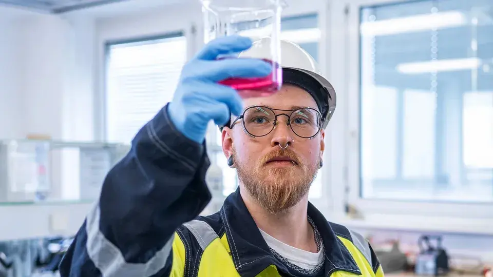 A lab technician wearing a hard hat, safety glasses, and gloves holds up a beaker containing a pink liquid for inspection. The scene takes place in a bright laboratory, emphasizing the focus on scientific analysis and safety in an industrial or research setting.