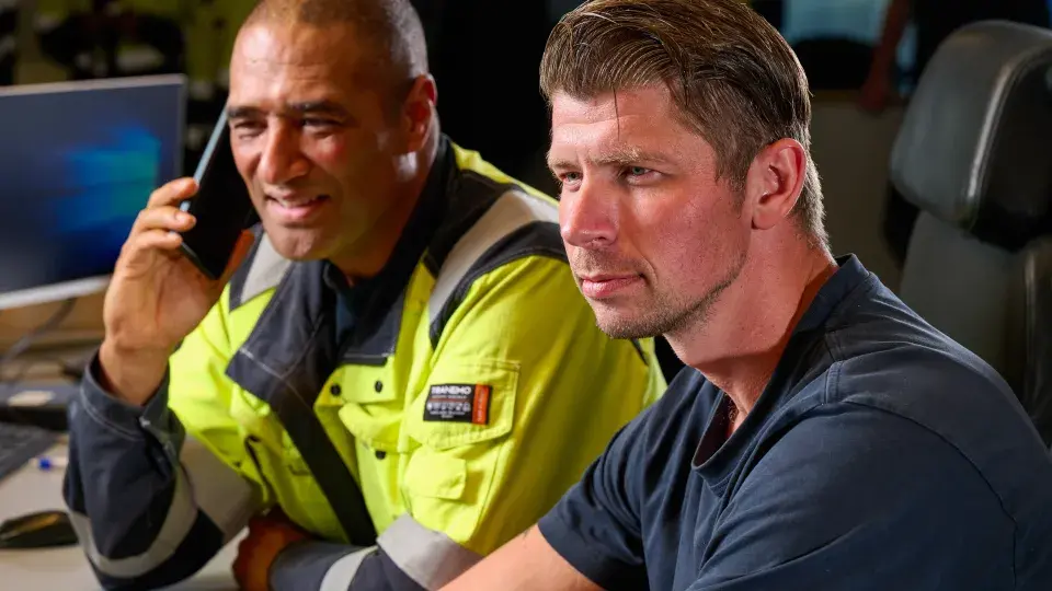 Two men in a control room, one wearing a high-visibility jacket and talking on the phone, while the other looks intently at a monitor.