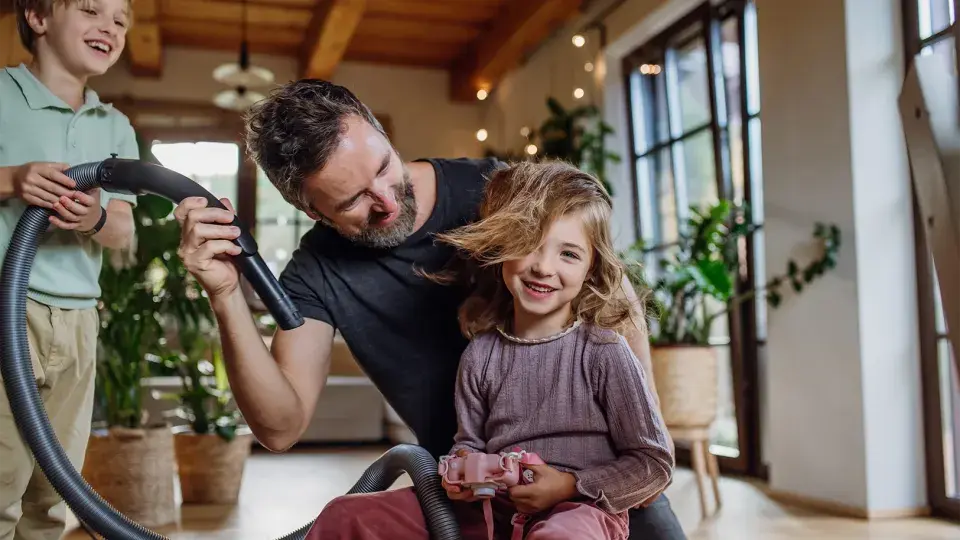 A father playfully using a vacuum hose to blow air into his daughter's hair while his son laughs nearby, creating a fun family moment indoors.