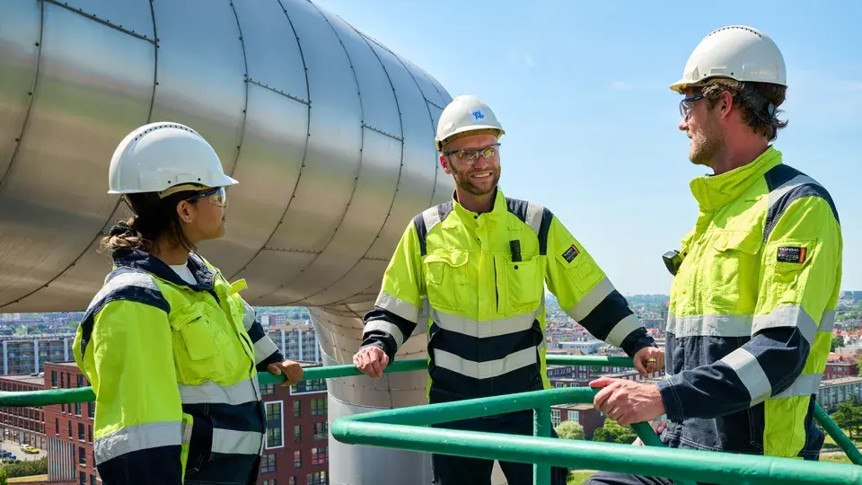 Three Uniper workers wearing high-visibility clothing and safety helmets standing outdoors on a platform near large industrial equipment, engaged in conversation.
