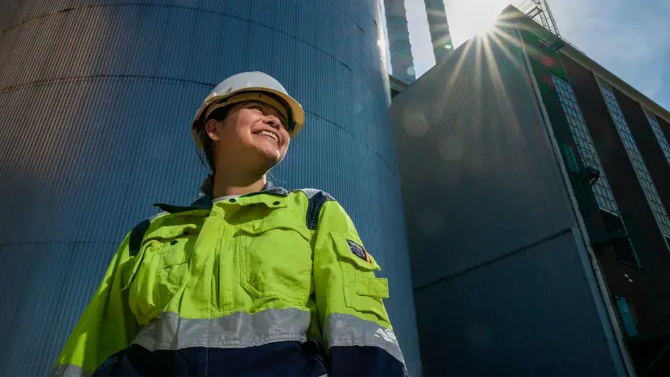 A worker in a high-visibility jacket and hard hat smiling while standing outside near industrial silos, with sunlight shining behind them.