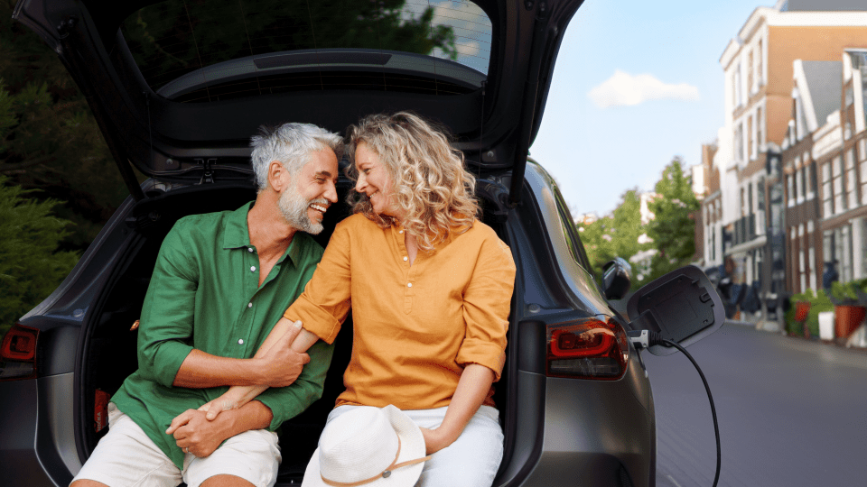 Smiling middle-aged couple sitting at the open trunk of an electric car, with a charging cable connected, sharing a warm moment in a cozy, urban setting.