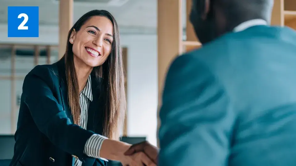 A smiling woman in a professional setting shaking hands with another person, with a blue box containing the number '2' in the top left corner.