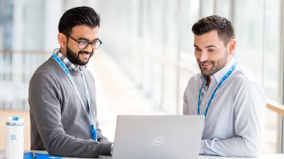 Two male colleagues sitting at a table, smiling while working together on a laptop in a bright office space, with a notebook and a Uniper-branded water bottle on the table