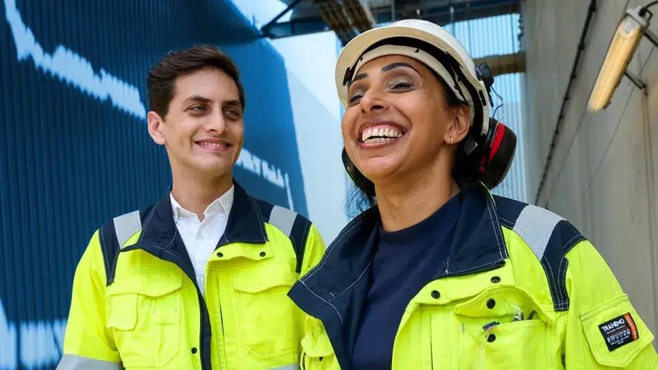Two workers in safety gear, one wearing a helmet and ear protection, smiling while standing in an industrial setting.