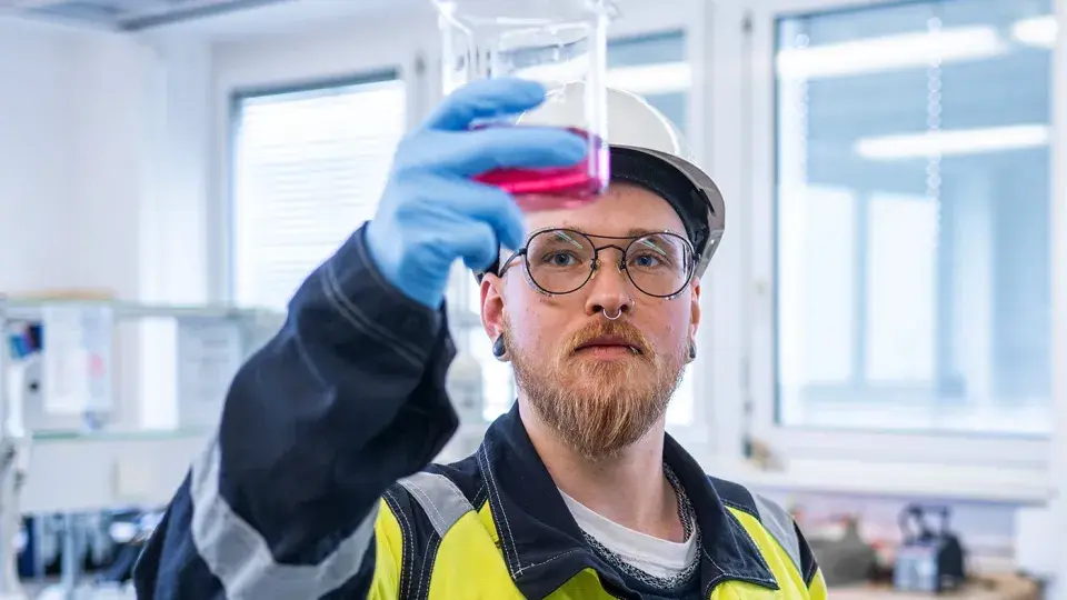 A technician in safety gear and a hard hat holds up a beaker with a pink liquid, carefully inspecting it in a laboratory setting.