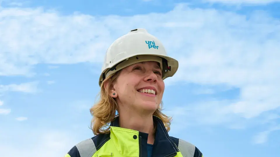 A smiling worker wearing a Uniper-branded hard hat and high-visibility jacket, standing outdoors against a bright blue sky.