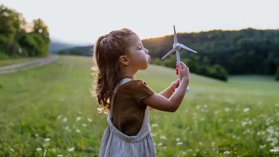 A young girl standing in a field at sunset, holding a toy wind turbine and blowing on it, with trees and hills in the background.