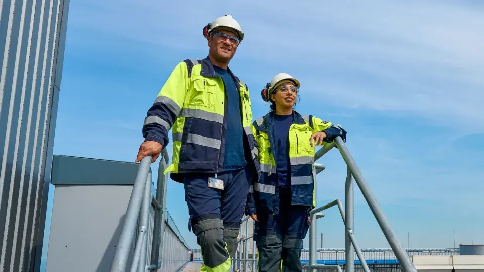 Two workers in high-visibility safety jackets, hard hats, and protective glasses standing on an outdoor industrial platform under a clear sky.