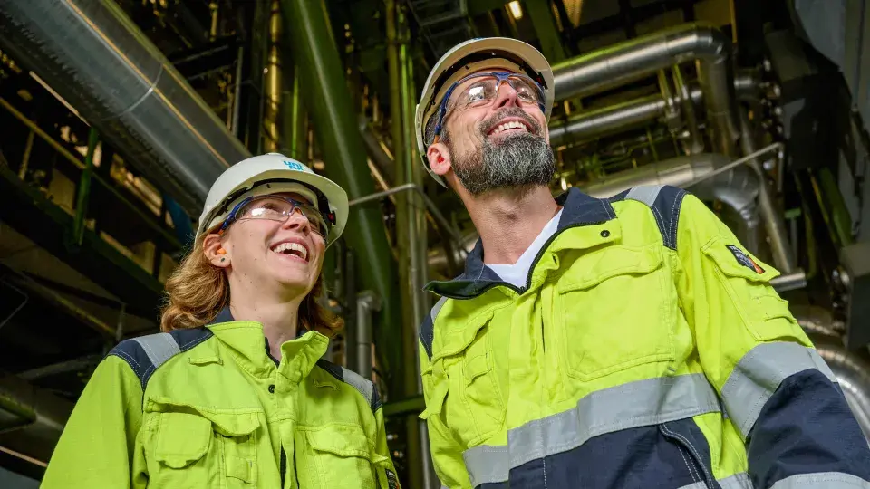 Two workers in high-visibility safety jackets, hard hats, and protective glasses smiling while standing in an industrial setting, with metal pipes and machinery in the background.