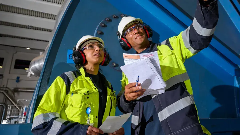 Two industrial workers in high-visibility jackets, hard hats, and ear protection reviewing documents and taking notes while inspecting machinery inside a facility.